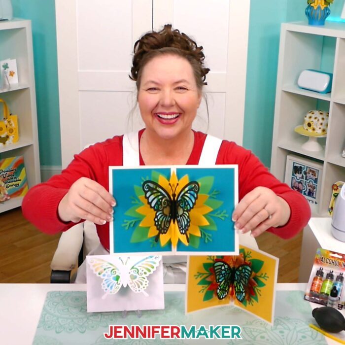 Jennifer Maker smiles at her craft table and shows off Alcohol Ink On Paper butterfly pop-up cards she made in her tutorial