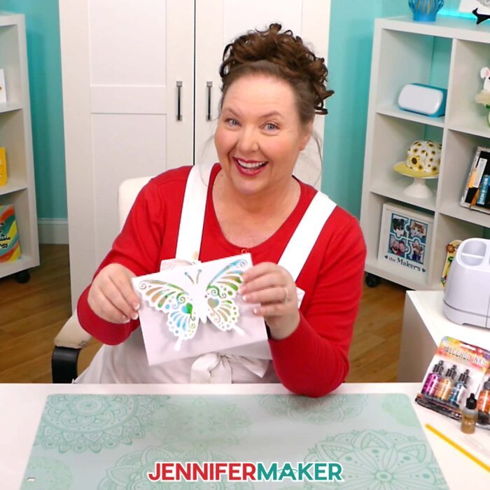 Jennifer Maker smiles at her craft table and shows off Alcohol Ink On Paper butterfly pop-up cards she made in her tutorial