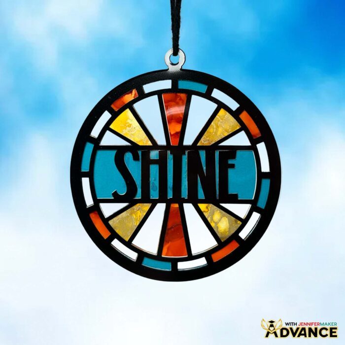 An Alcohol Ink Paper Stained Glass window project featuring text reading "SHINE" and cheerful blue, yellow, and orange colors with black "solder" lines cut from vinyl hangs against a cloudy blue sky 