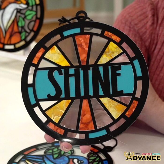 Hands hold a Alcohol Ink Paper Stained Glass window project featuring text reading "SHINE" and cheerful blue, yellow, and orange colors with black "solder" lines cut from vinyl