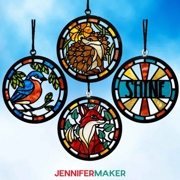 Four Alcohol Ink Paper Stained Glass window projects featuring a bird, hedgehog, "shine," and fox designs, hanging against a bright blue sky with fluffy clouds