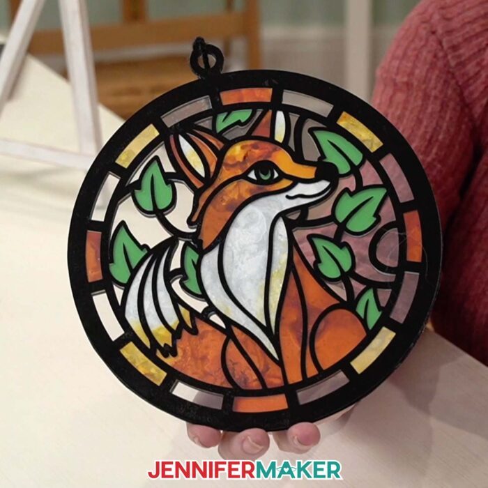 Hands hold an Alcohol Ink Paper Stained Glass window project featuring a red and orange fox,  with black "solder" lines cut from vinyl and clear acetate to represent the glass