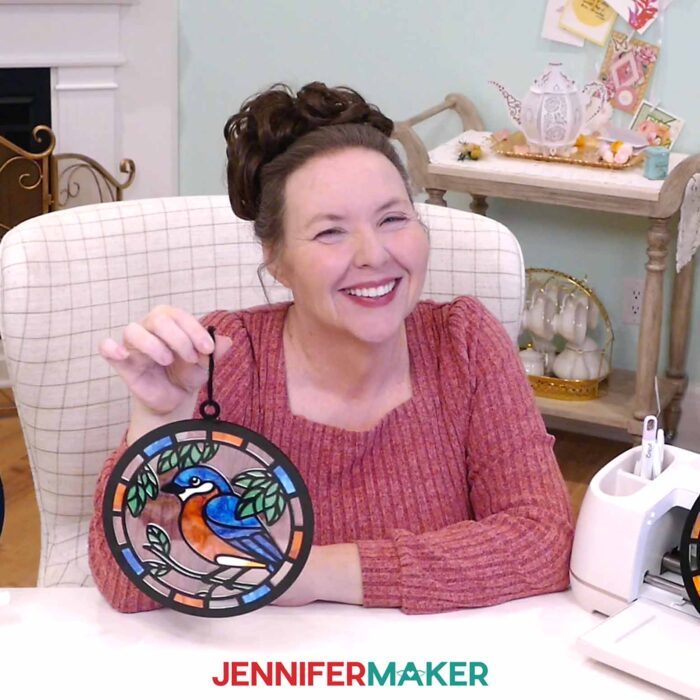 Jennifer Maker shows off an Alcohol Ink Paper Stained Glass window project featuring a colorful bluebird design in cheerful shades of blue, orange, and green with black "solder" lines cut from vinyl