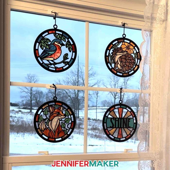 Four Alcohol Ink Paper Stained Glass window projects featuring a bird, hedgehog, "shine," and fox designs, hanging in a window looking out over a bright, snowy landscape 