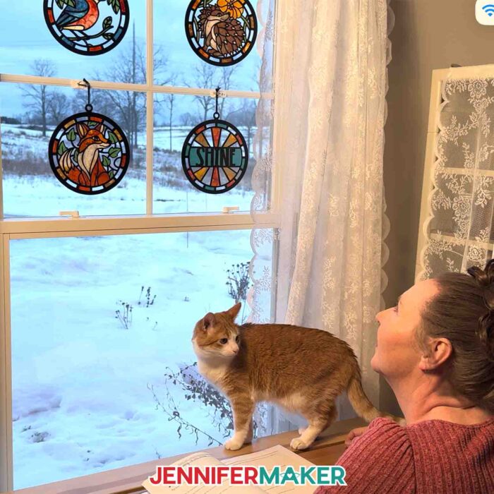 Jennifer Maker and her cat Butterscotch look at four Alcohol Ink Paper Stained Glass window projects featuring a bird, hedgehog, "shine," and fox designs, hanging in a window looking out over a snowy landscape