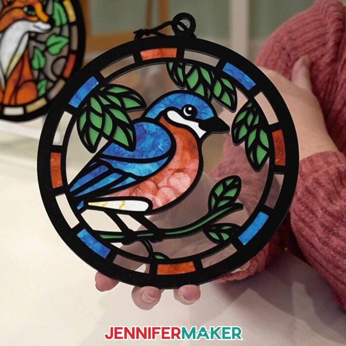 Hands hold a Alcohol Ink Paper Stained Glass window project featuring a bluebird,  with black "solder" lines cut from vinyl and clear acetate to represent the glass