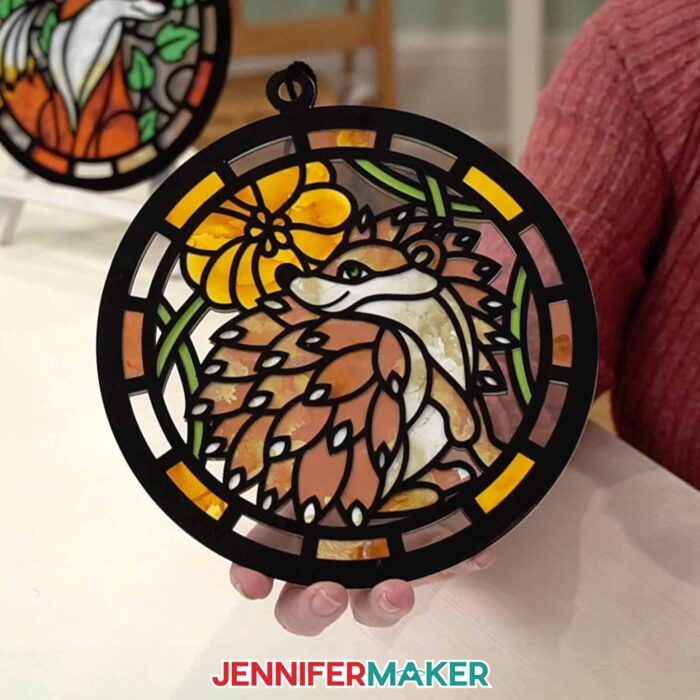 Hands hold a Alcohol Ink Paper Stained Glass window project featuring a brown hedgehog and a yellow flower,  with black "solder" lines cut from vinyl and clear acetate to represent the glass