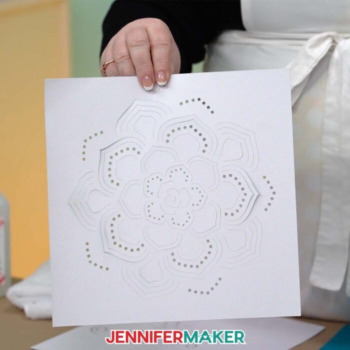 A hand holds a piece of white cardstock with a succulent design cut into it by a Cricut, ready to be assembled into a Backlit Shadow Box 