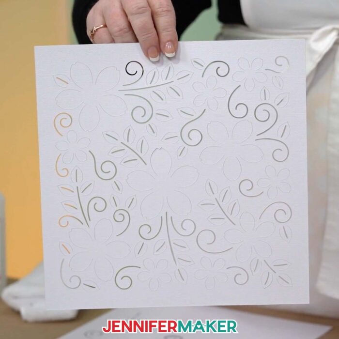 A hand holds up a 12 inch by 12 inch piece of white cardstock with flower, leaf, and swirl shapes cut into it by a Cricut cutting machine