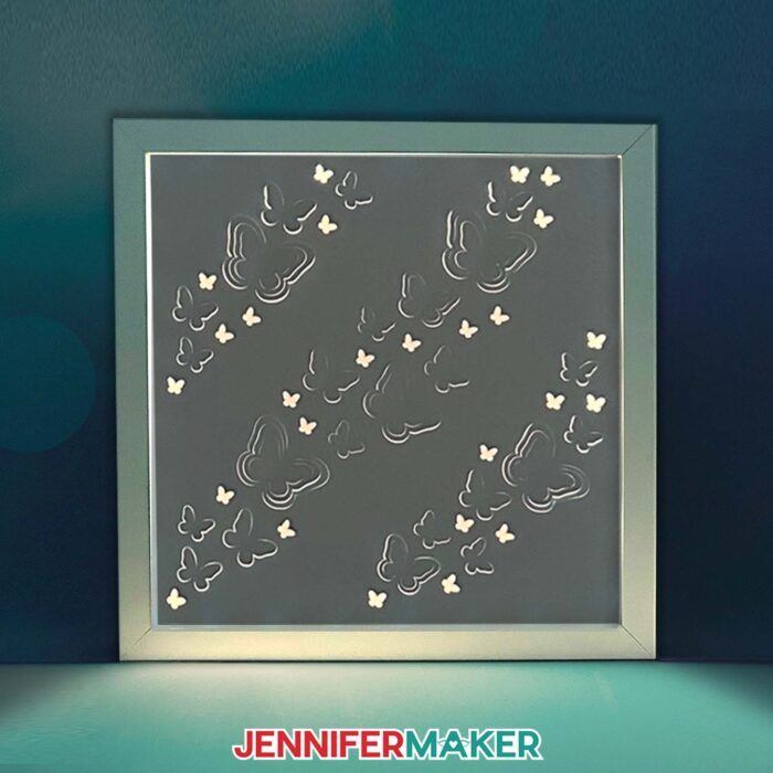 A Backlit Shadow Box project with butterfly cutouts running diagonally across it glows from in from inside a square white frame in a dimly lit space