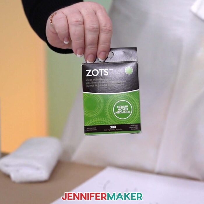 A hand holds a small green box of Zots medium-sized glue dots to be used for sticking the battery pack to the back of the Backlit Shadow Box frame