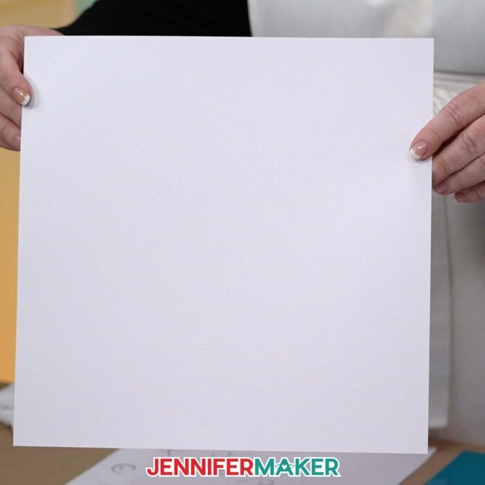 Hands hold up a sheet of 12 inch by 12 inch white cardstock that is used to make a Backlit Shadow Box