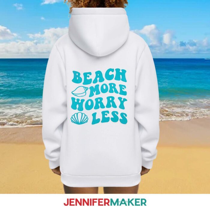 A person facing away from the camera wearing a white hoodie with a vinyl design on the back reading "beach more worry less" with a with blue seashells.