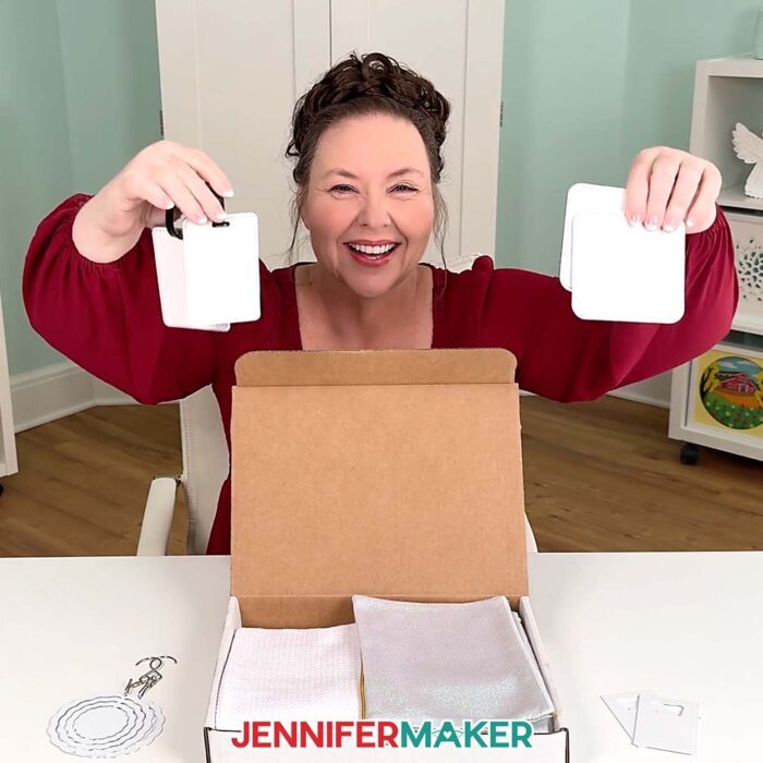 Jennifer smiles and holds up the luggage tag and coaster blanks from the Beginner Sublimation Kit with the box displayed one the table in front of her