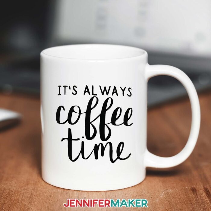 White mug on a wooden desk with a laptop in the background. The mug reads "It's Always Coffee Time" in black vinyl brush-script. Learn all about the best Cricut vinyl for coffee mugs in JenniferMaker's new tutorial!