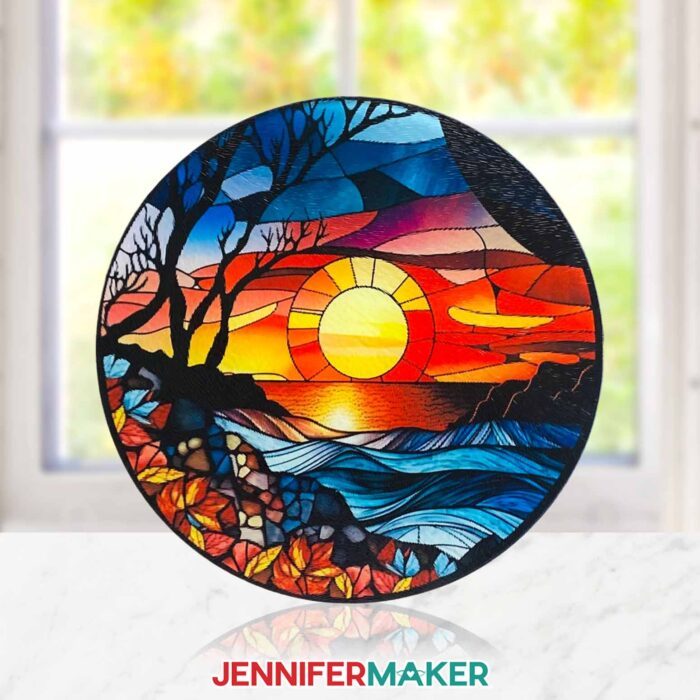 Can you sublimate on glass cutting boards? Learn how with JenniferMaker's tutorial! A round stained glass cutting board featuring a colorful sunset scene sits propped up in front of a sunny window.