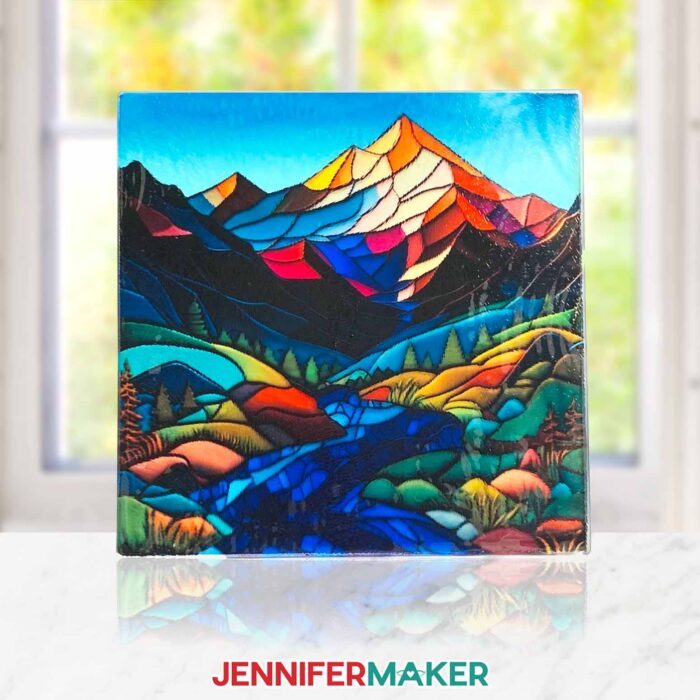 Can you sublimate on glass cutting boards? Learn how with JenniferMaker's tutorial! A square stained glass cutting board featuring a colorful mountain scene sits propped up in front of a sunny window.
