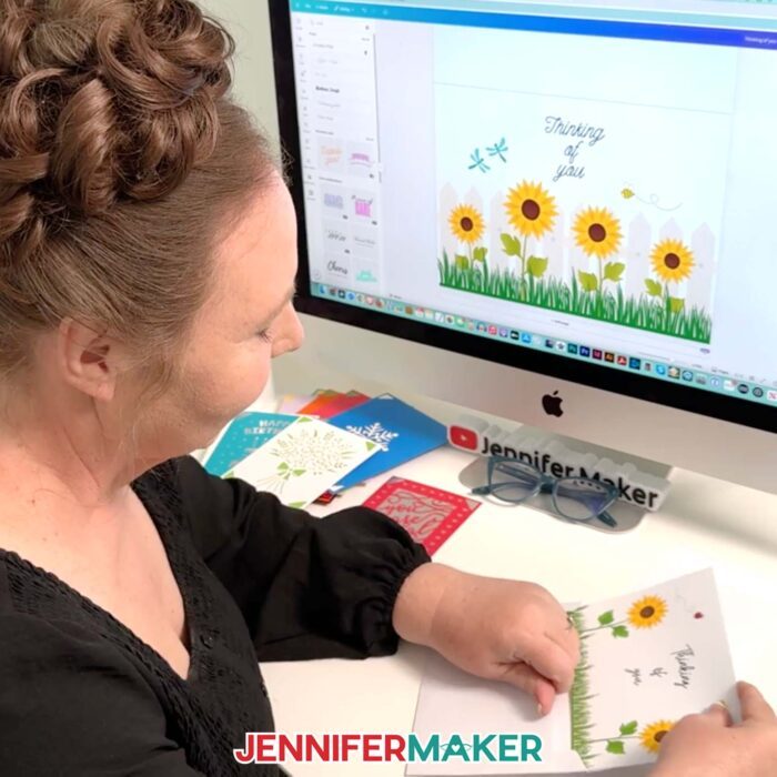 Jennifer Maker sits at her computer desk with Canva on the screen, and she's putting a card she made -- a folding card with sunflowers, a bee, a ladybug, and "Thinking Of You" printed on the front -- into a white envelope. Learn to make your own with the How To Make Cards In Canva tutorial