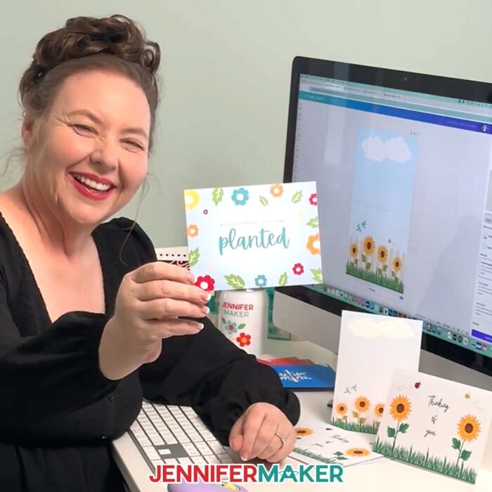Jennifer Maker sits at her computer desk with Canva on the screen, and she's holding a card she made -- a card with multicolored flowers and "Bloom Where You Are Planted" printed on the front. Learn to make your own with the How To Make Cards In Canva tutorial