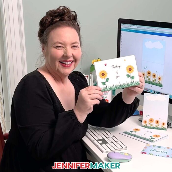Jennifer Maker sits at her computer desk with Canva on the screen, and she's holding a card she made -- a folding card with sunflowers, a bee, a ladybug, and "Thinking Of You" printed on the front. Learn to make your own with the How To Make Cards In Canva tutorial