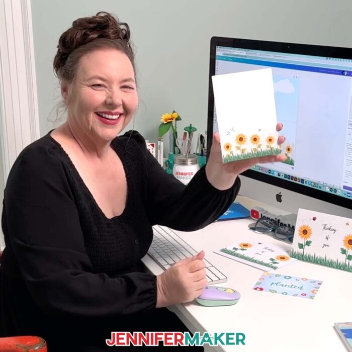 Jennifer Maker sits at her computer desk with Canva on the screen, and she's holding a card she made -- a note card with sunflowers printed on the front. Learn to make your own with the How To Make Cards In Canva tutorial