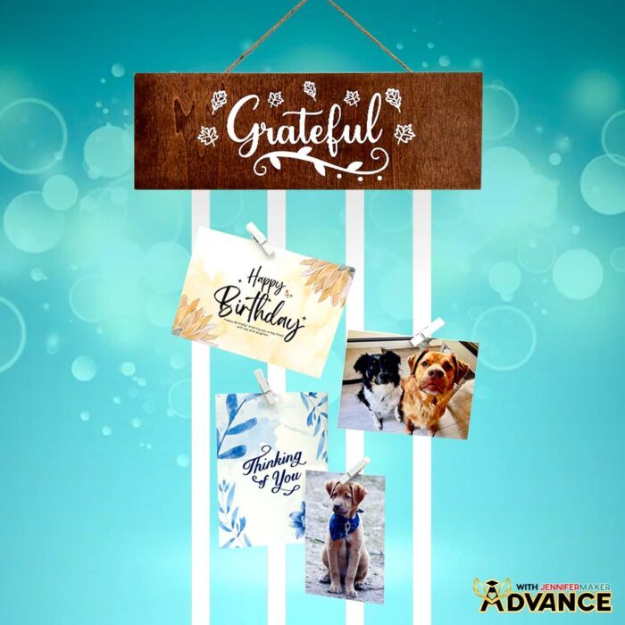 Custom wooden display panel for an interchangeable ribbon greeting card hanger. White vinyl that reads "Grateful" surrounded by leaf decals on a faux-stained brown panel with greeting cards attached to ribbons dangling from the panel.