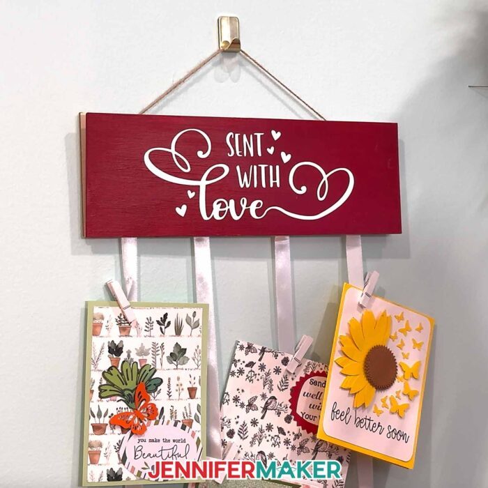 An greeting card holder hangs on a wall with a red and white "Sent With Love" display panel showing and cards clipped to the holder's ribbons.