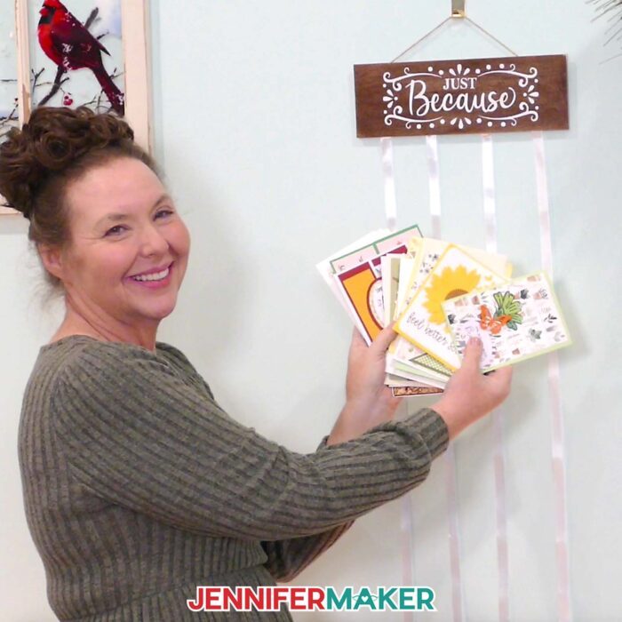 Jennifer holds up a pile of greeting cards next to a hanging card holder with a display panel that says "Just Because."