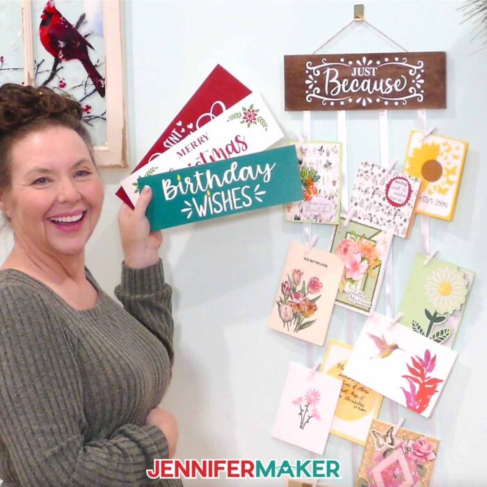 Jennifer holds up three painted display panels beside a "Just Because" hanger on the wall with greeting cards attached to it.