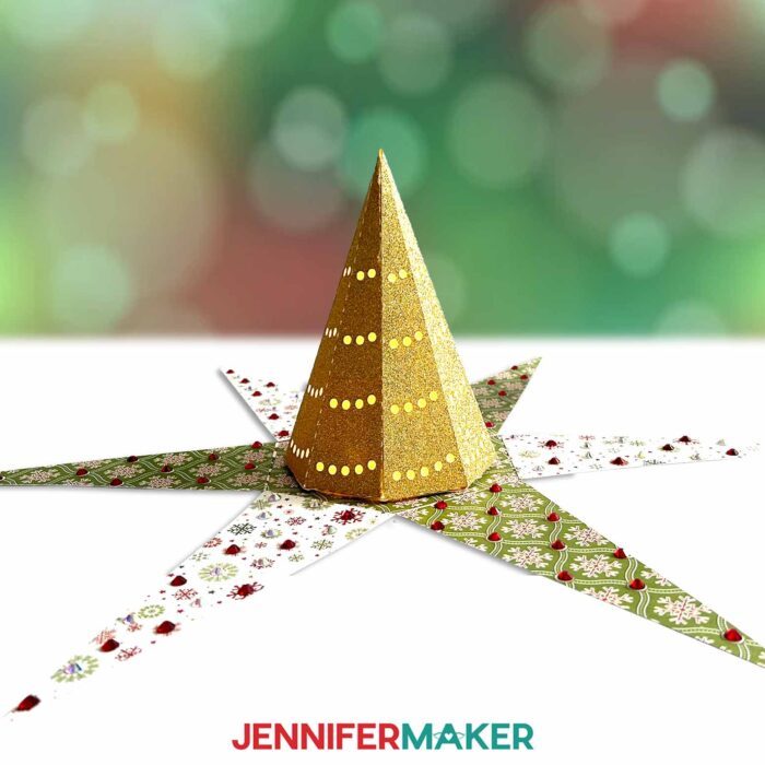 A Christmas Tree Explosion Box with patterned holiday paper sits, open, on a white surface, revealing the smaller glittery gold tree inside