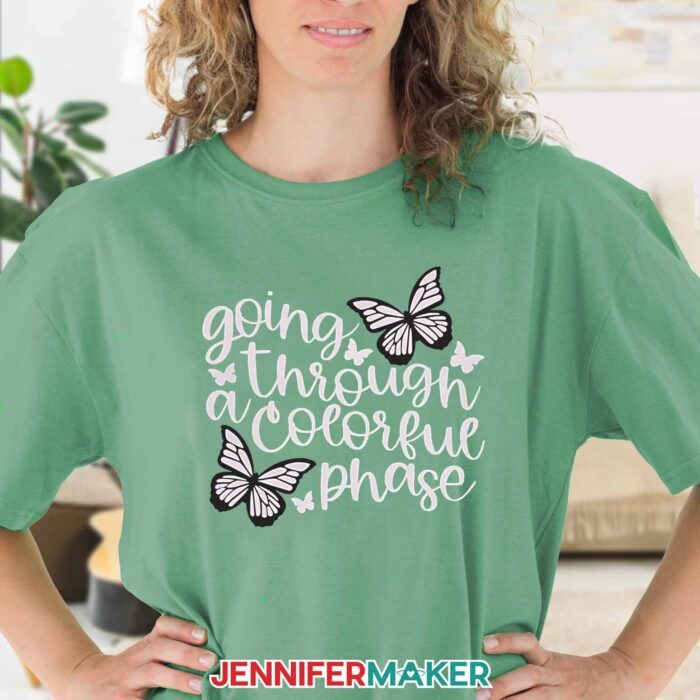 Model wearing a green shirt with a UV reactive color changing design on it with butterflies that says "Going Through a Colorful Phase". Learn to use color changing HTV with JenniferMaker's tutorial!