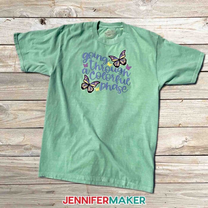 Green t-shirt with UV reactive color changing design on it with butterflies that says "Going Through a Colorful Phase". Learn to use color changing HTV with JenniferMaker's tutorial!