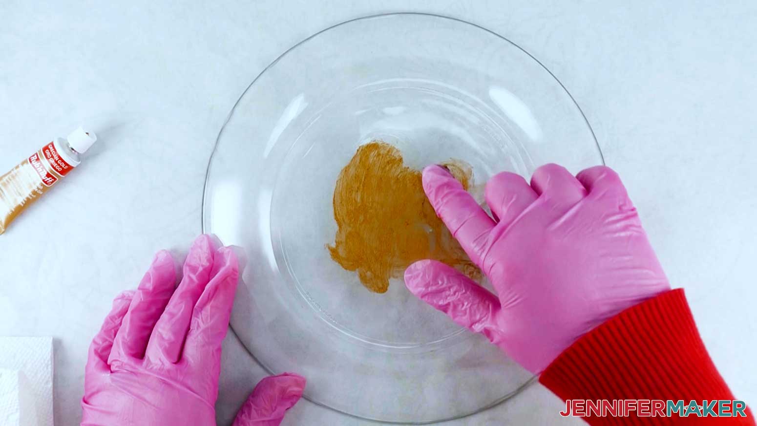 An overhead photo showing gold Rub n Buff paint being applied to a clear plate with a finger wearing a pink glove. Another gloved hand holds the plate in place, and a tube of Rub n Buff paint and a paper towel are displayed on the left.