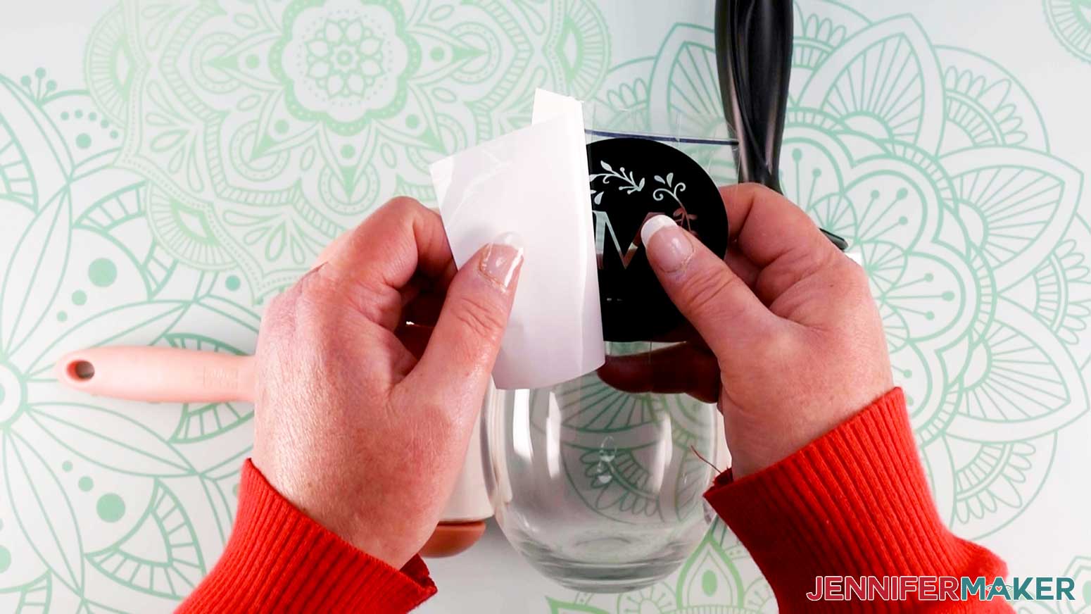 An overhead photo showing one hand removing the paper backing from the weeded vinyl design for the color glass etching project, with a clear wine glass positioned on the work surface below