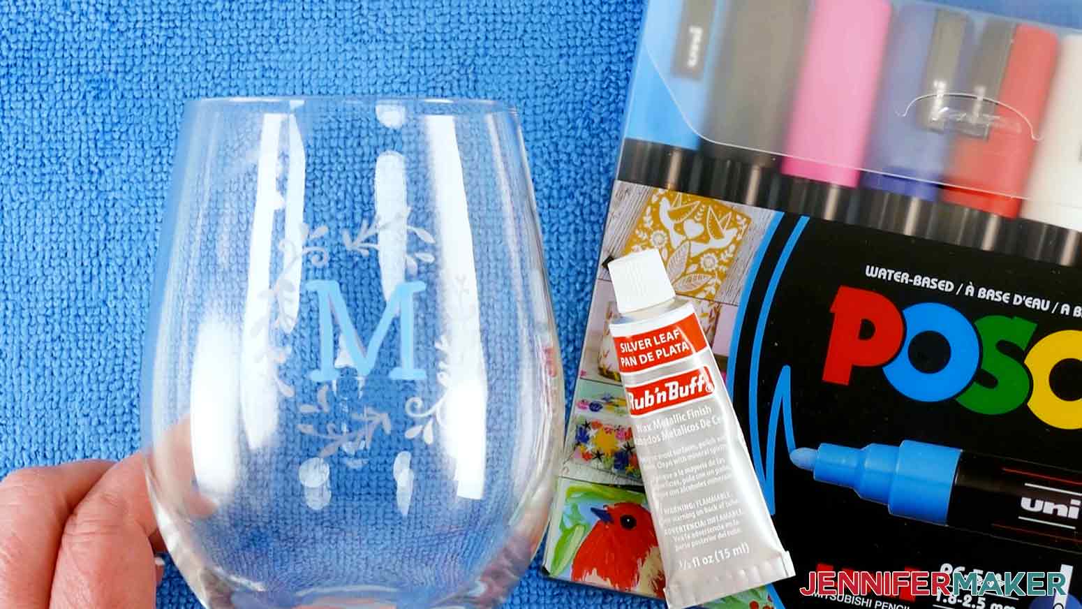 An overhead photo showing a finished color glass etching wine glass project with a silver colored floral design surrounding a blue M. A package of water-based paint markers and a tube of silver leaf Rub n Buff is displayed to the right of the glass.