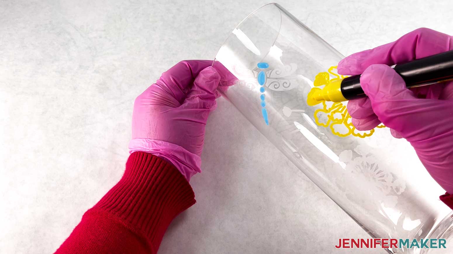 An overhead photo showing a gloved hand holding a vase in place while another gloved hand paints an etched design with a yellow paint marker. The dragonfly part of the color glass etching design is painted in blue.