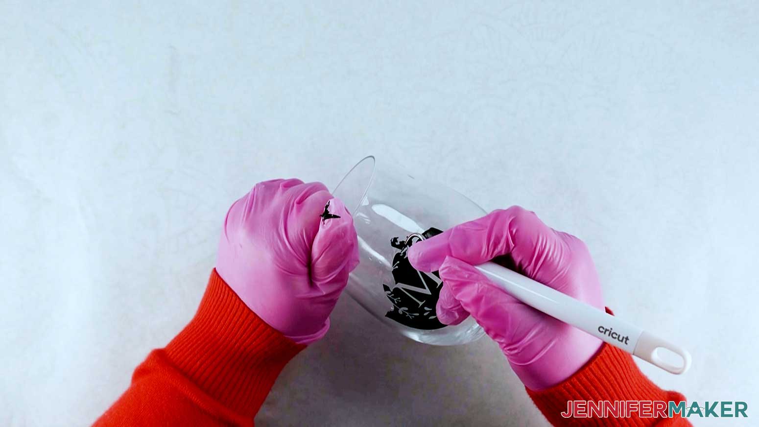 An overhead photo showing a gloved hand holding a wine glass in place while the other gloved hand uses a weeding tool to remove small pieces of vinyl to reveal the etched design on the glass