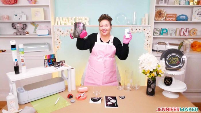 Jennifer Maker wearing a pink apron, and holding a respirator and a bottle of Armour Etch for color glass etching