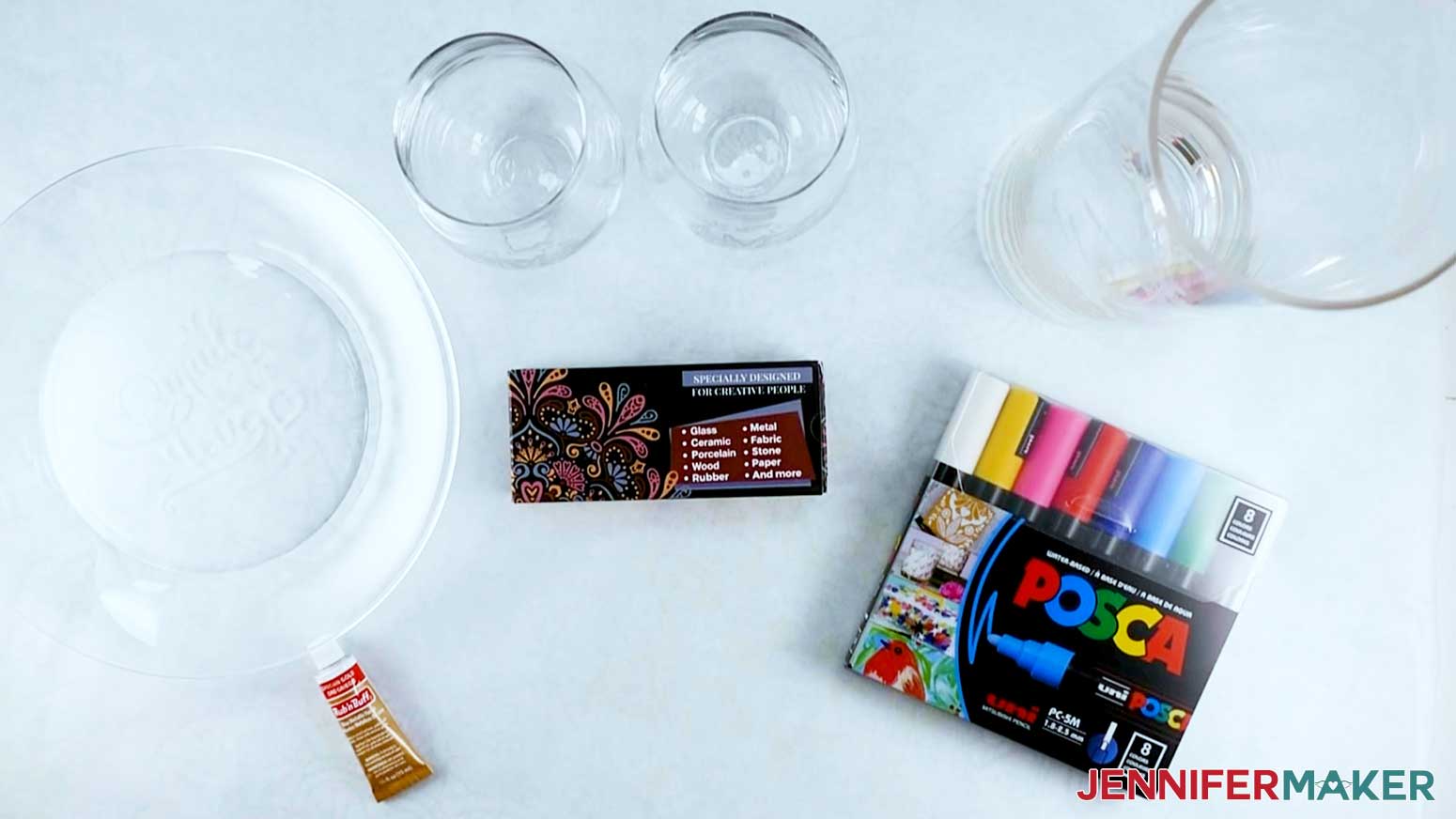 An overhead photo of a glass plate with a tube of gold Rub n Buff displayed below it, two stemless wine glasses with a package of oil-based paint pens displayed below them, and a glass vase with a package of water-based paint markers displayed below it