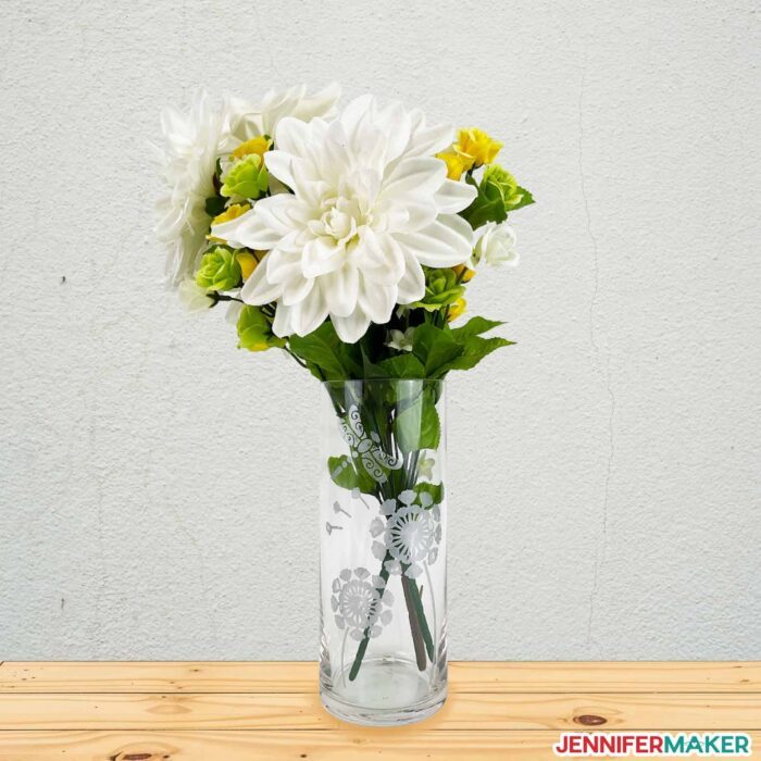 A clear glass vase with a floral white color etching design holding white flowers and sitting on a table.