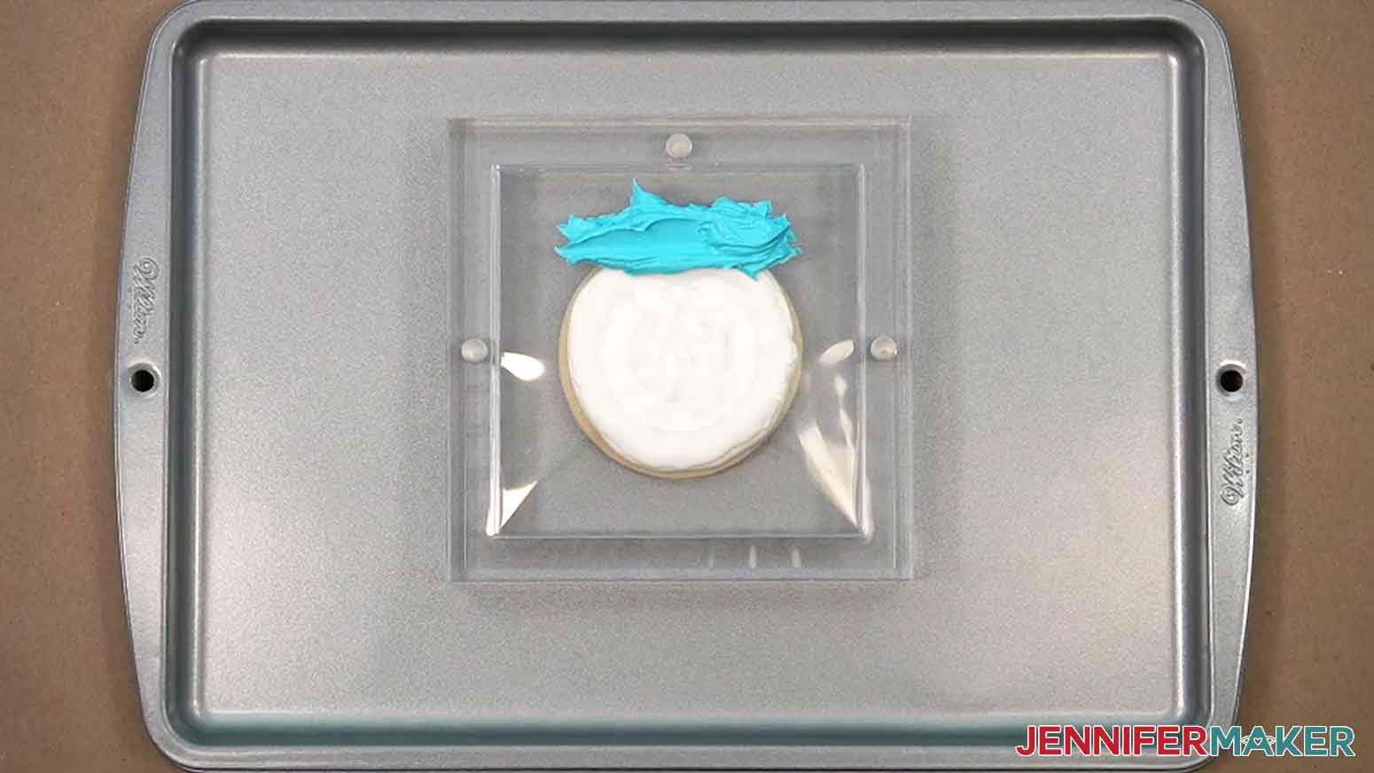 The iced cookie has been placed under the cookie stencil holder and a line of thicker icing is shown above the cutouts on the stencil.