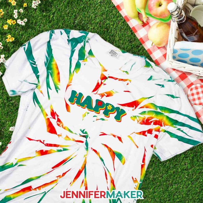 White sublimation tie dye shirt with multicolored swirl designs and a Happy illustrated design laid on a picnic scene.