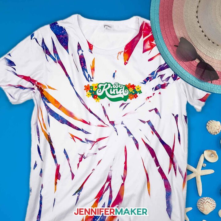 White sublimation tie dye shirt with multicolored swirl designs and a Be Kind illustrated design laid on a beach scene.