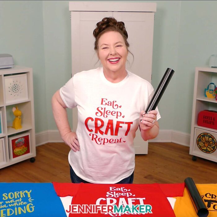 Jennifer Maker wears a white T-shirt she decorated with iron-on vinyl to say "Eat, Sleep, Craft, Repeat," and holds a roll of black HTV. Learn how to make your own in the Cricut Iron-On Vinyl Tutorial