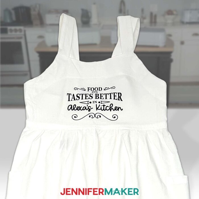 A white apron decorated with a black HTV sentiment "Food Tastes Better in Alexa's Kitchen" made from bonus materials included with the new Cricut Maker 4 & Cricut Explore 4!