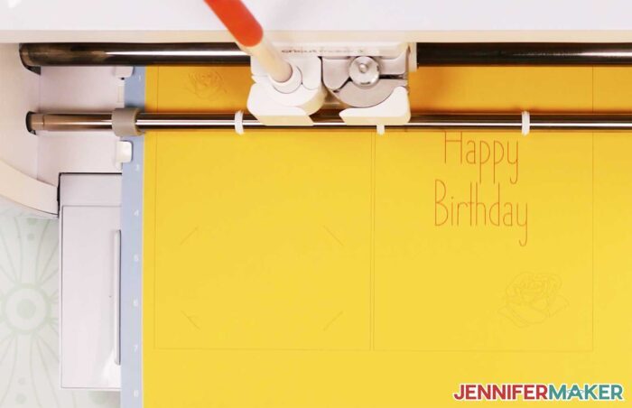 A Cricut machine writing Happy Birthday on a yellow paper using an orange pen in one of its clamps for the Cricut Writing Fonts tutorial.
