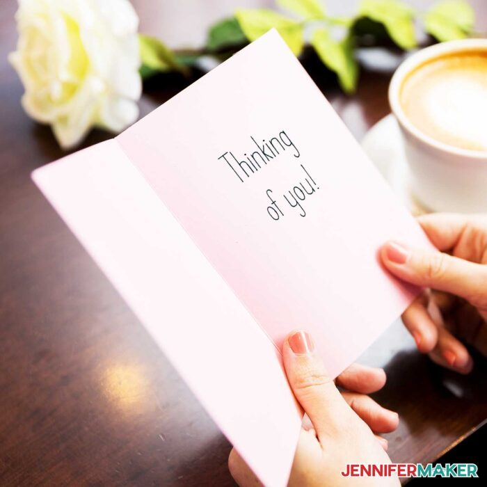 Hands holding a white paper card with "Thinking of You" written using Cricut writing font options.