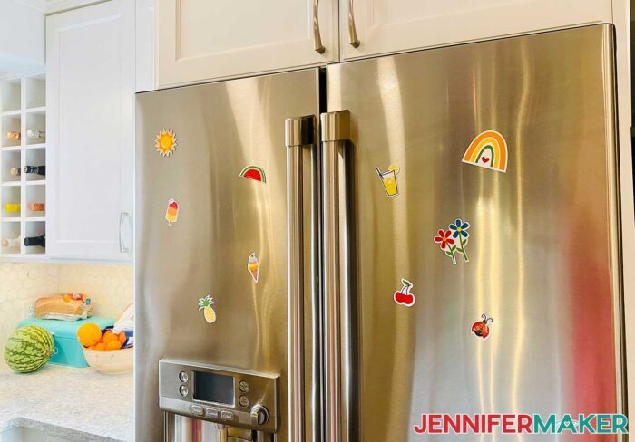 DIY fridge magnets with bright designs of fruits, summer snacks, and a ladybug on a stainless steel refrigerator.