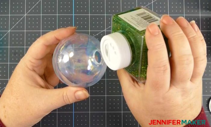 Pouring green glitter into a clear ornament to make DIY glitter ornaments