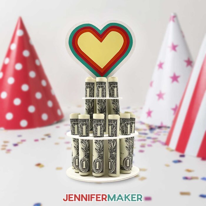 Colorful money cake decoration holding rolled dollar bills a in two-tier column, topped with a multicolor heart, made using a JenniferMaker SVG file.